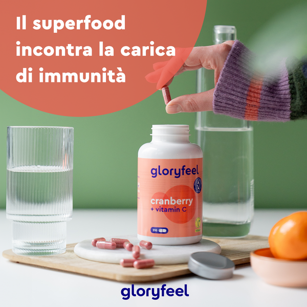 Una mano tiene una capsula GloryFeel GmbH Italy Cranberry + Vitamina C sopra una bottiglia aperta su un tavolo con acqua, un bicchiere e un&