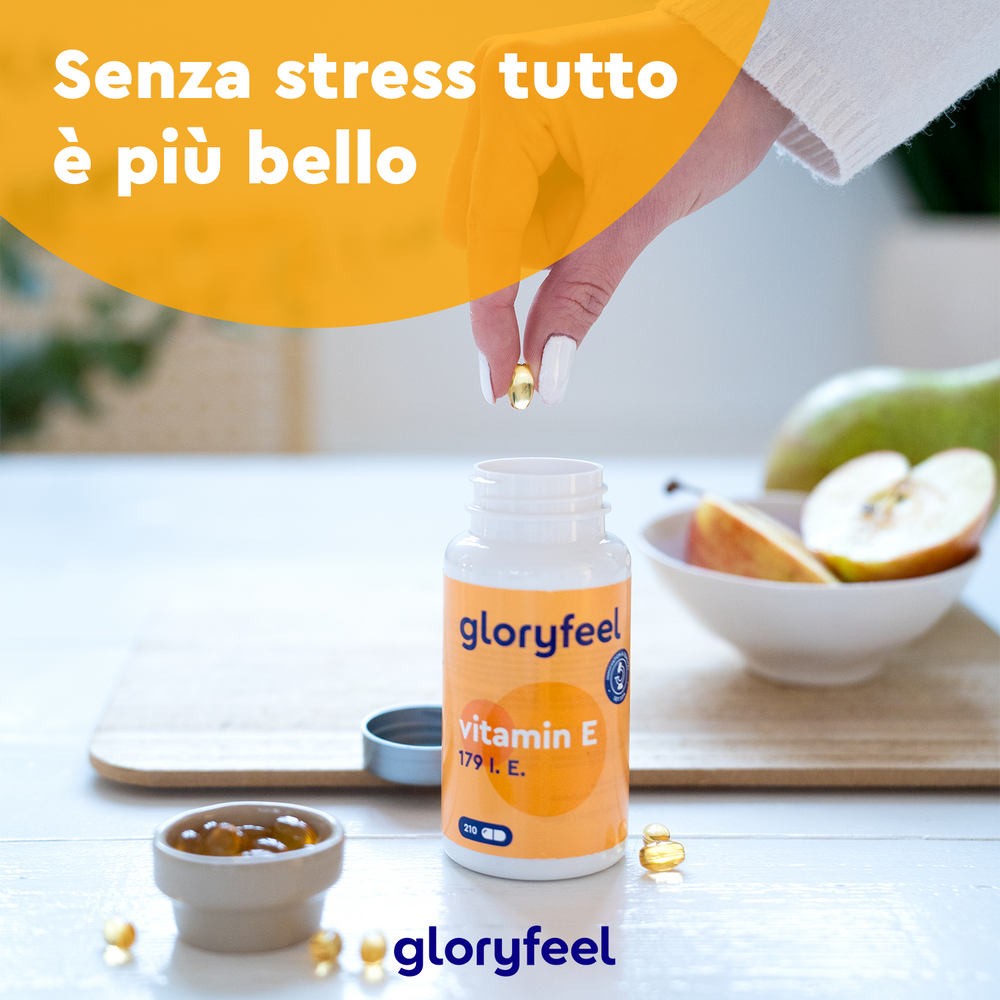 Una mano afferra una capsula da una bottiglia di Vitamina E Softgel di gloryfeel Italia, posta accanto a una ciotola di fette di mela e a un&
