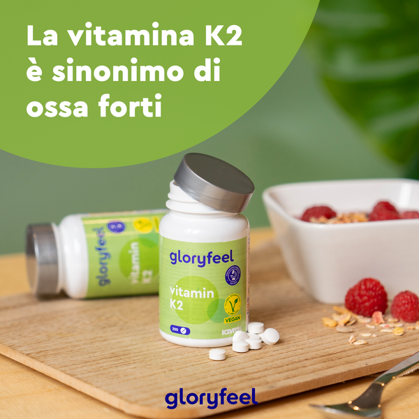 Due flaconi di gloryfeel Italia Vitamina K2 sono appoggiati su una superficie di legno, uno aperto con pillole sparse. Una ciotola piena di lamponi, avena e noci completa la scena sotto il testo italiano, promuovendo naturalmente ossa più forti e una vita sana.