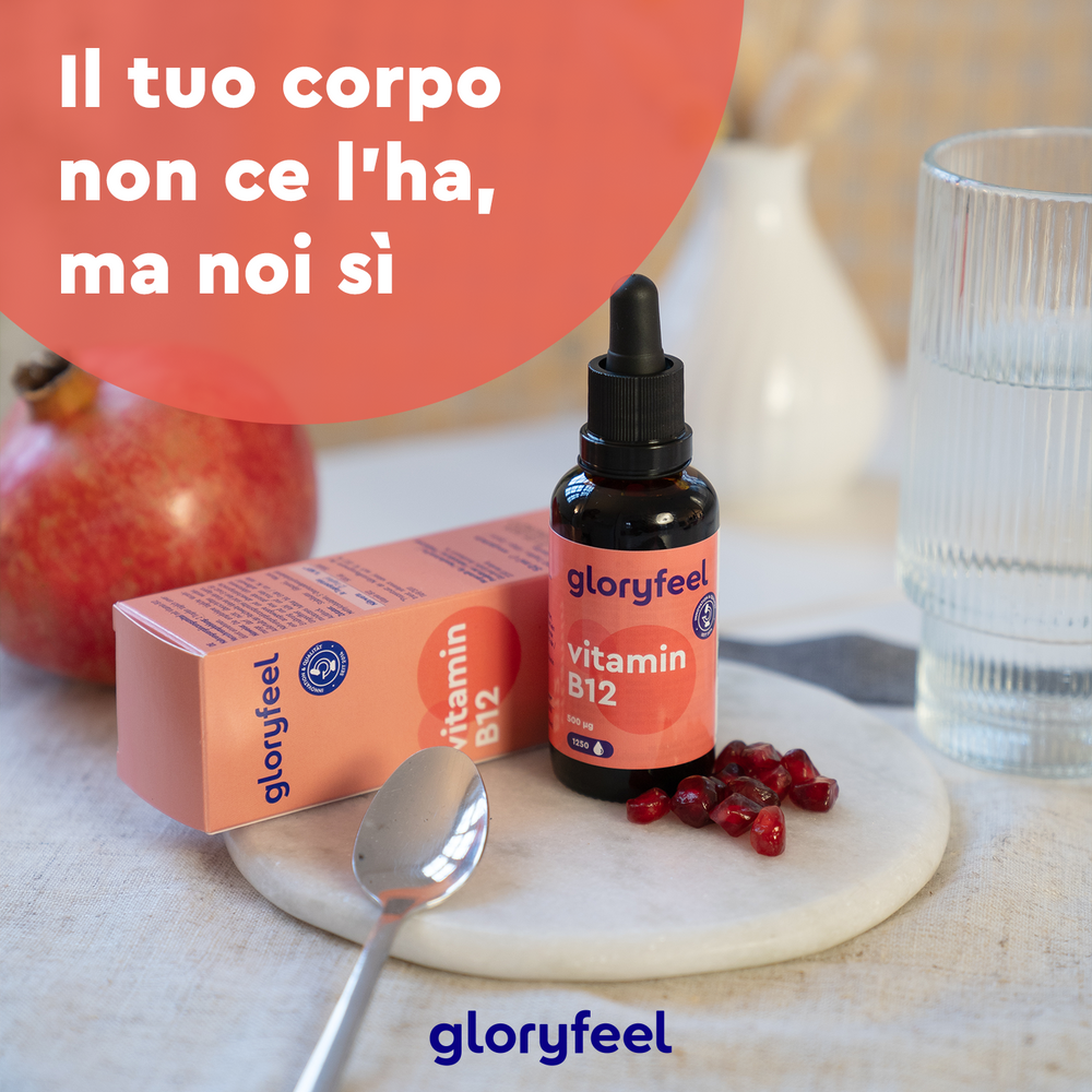 La bottiglia e la scatola di GloryFeel GmbH Italy Vitamina B12 Gocce sono su un tavolo con un cucchiaio, semi di melograno vibranti, un frutto intero e un bicchiere d&