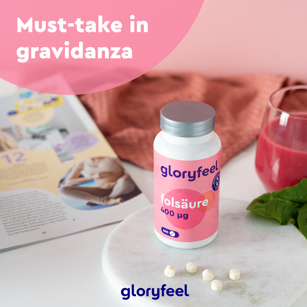 Una bottiglia di compresse di GloryFeel GmbH Italy Acido Folico è appoggiata su un tavolo con compresse sparse accanto a una rivista, un bicchiere di bevanda rossa e foglie di spinaci. Il testo rosa sopra recita &quot;Integratore gravidanza must-take&quot;, sottolineando il ruolo fondamentale della vitamina B9 in gravidanza.
