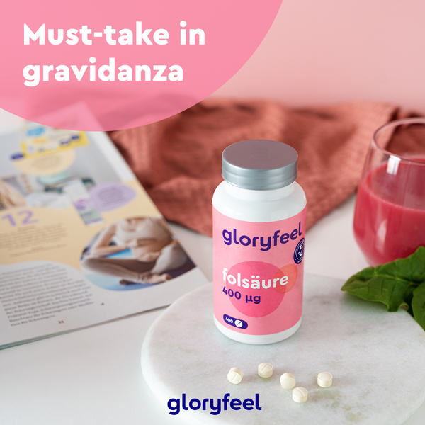 Una bottiglia di compresse di GloryFeel GmbH Italy Acido Folico è appoggiata su un tavolo con compresse sparse accanto a una rivista, un bicchiere di bevanda rossa e foglie di spinaci. Il testo rosa sopra recita &quot;Integratore gravidanza must-take&quot;, sottolineando il ruolo fondamentale della vitamina B9 in gravidanza.