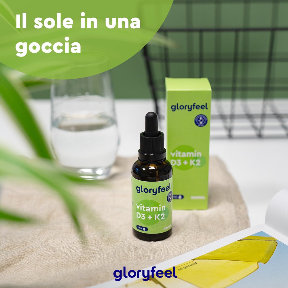 Una scatola e una bottiglia di Gloryfeel Italia Vitamina D3 K2 Gocce sono appoggiate su un telo beige, a simboleggiare una &quot;dieta equilibrata&quot;. Dietro di loro, su una superficie bianca, sono appoggiati un bicchiere d&