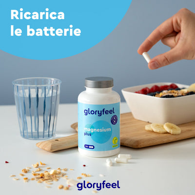 Una mano tiene una pillola su una tavola di legno con una bottiglia di Magnesio Plus 180 di GloryFeel GmbH Italia, un bicchiere d'acqua e una fruttiera. La scritta in italiano "Ricarica le batterie" promette una maggiore biodisponibilità.